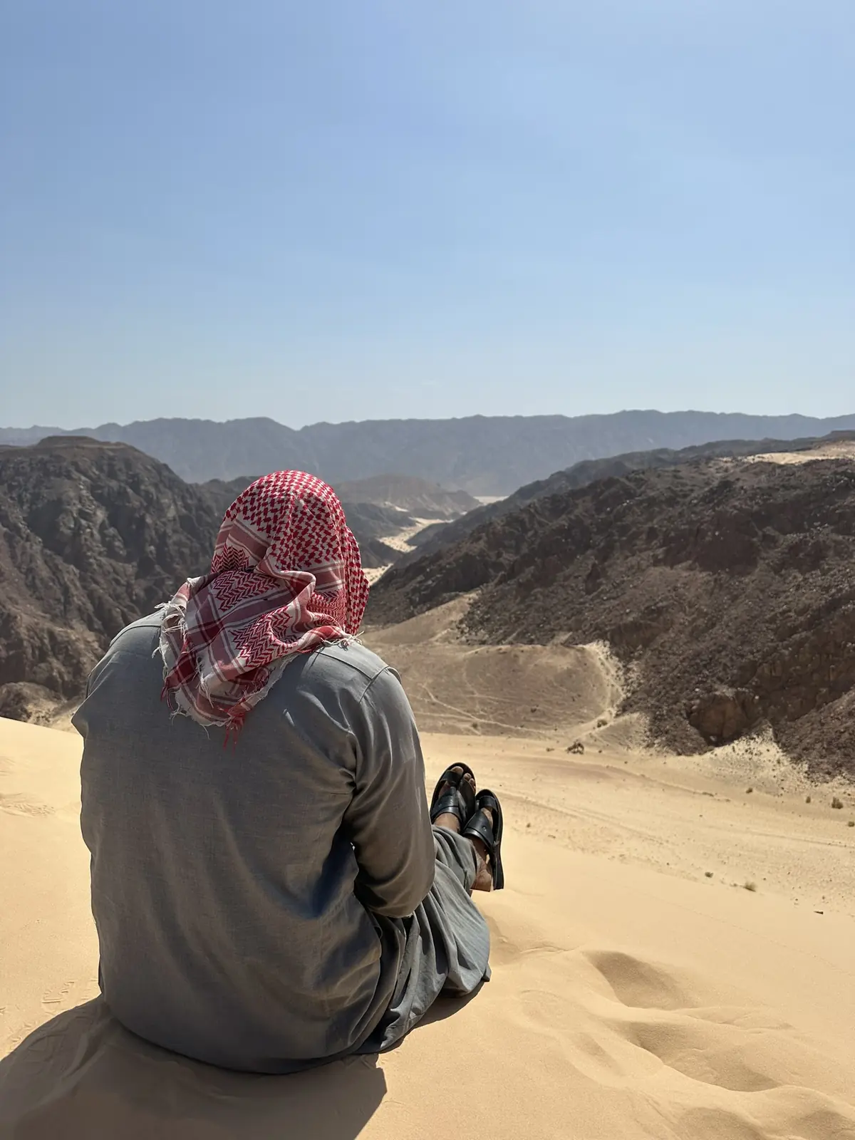 Beduine mit rot-weißer Kufiya sitzt auf einer Sanddüne und blickt über die Felslandschaft des Süd-Sinai