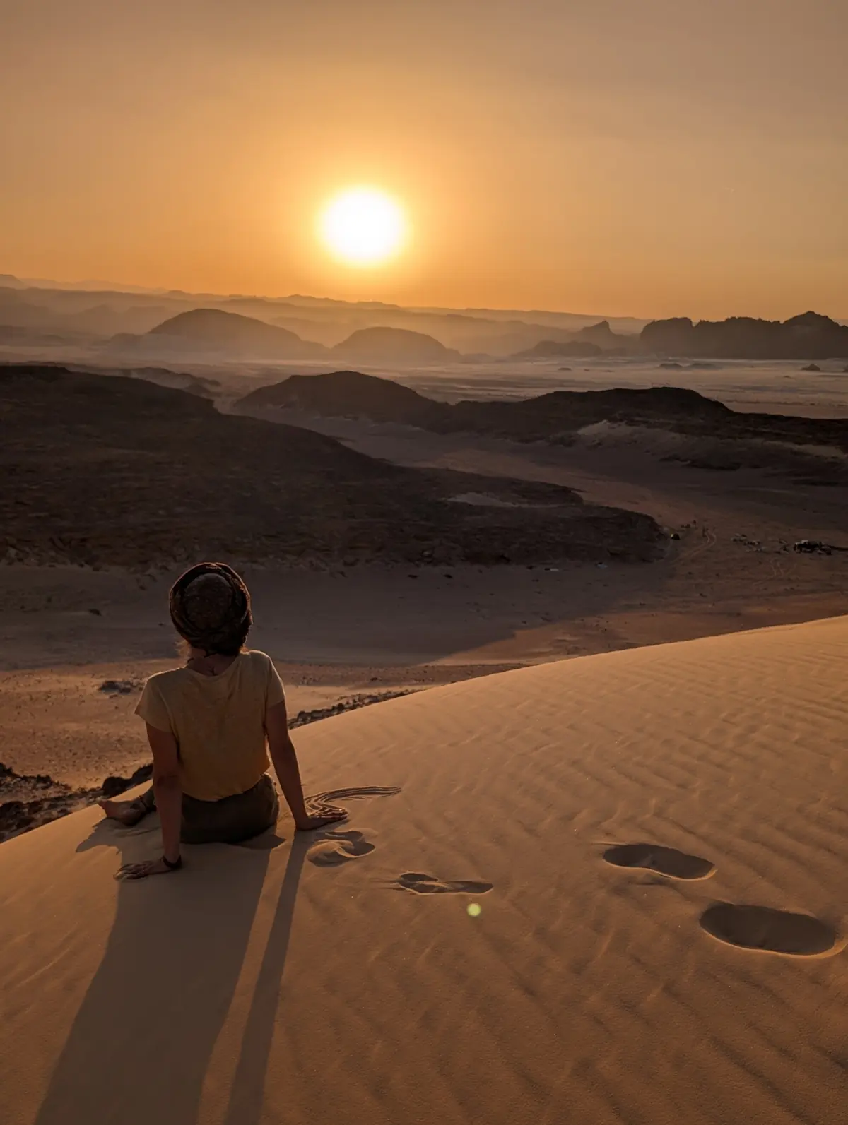Frau sitzt auf einer Sanddüne und blickt in den Sonnenuntergang über den Bergen des Süd-Sinai