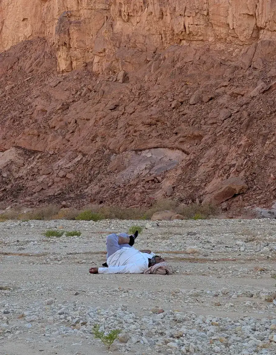 Beduine ruht ausgestreckt auf dem Wüstenboden vor einer rot leuchtenden Sandsteinwand im Süd-Sinai