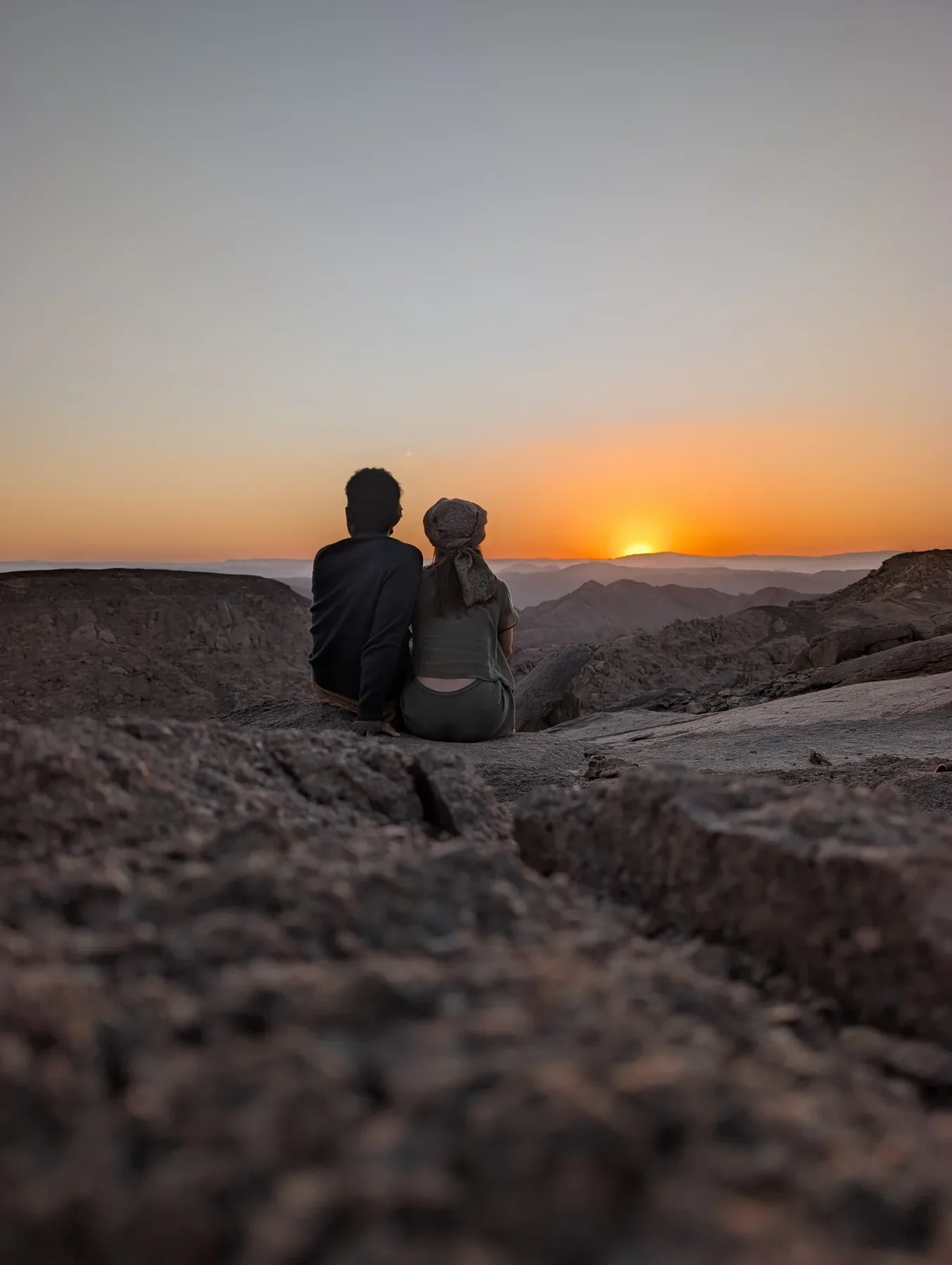 Zwei Menschen sitzen eng beieinander auf einem Felsen und blicken auf den Sonnenuntergang über den Bergen des Süd-Sinai